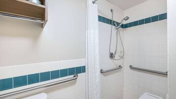 Une salle de bain avec des carreaux blancs et turquoise, une douche équipée d’un pommeau de douche à main, des toilettes dotées d’une barre d’appui et une étagère au-dessus des toilettes.