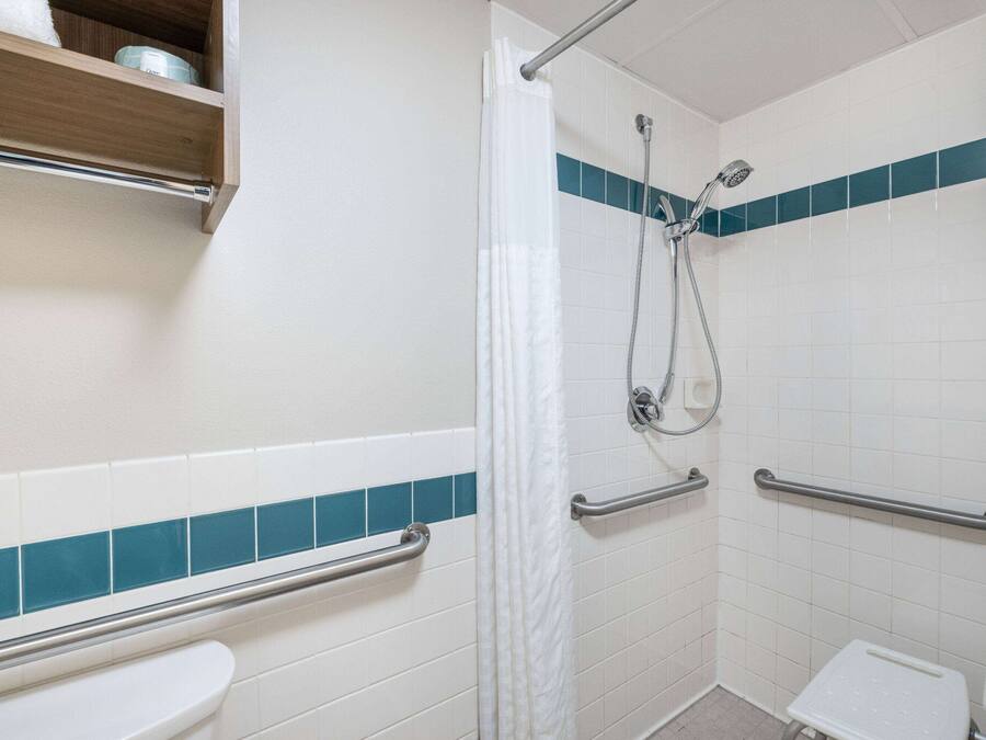 Une salle de bain avec des carreaux blancs et turquoise, une douche équipée d’un pommeau de douche à main, des toilettes dotées d’une barre d’appui et une étagère au-dessus des toilettes.
