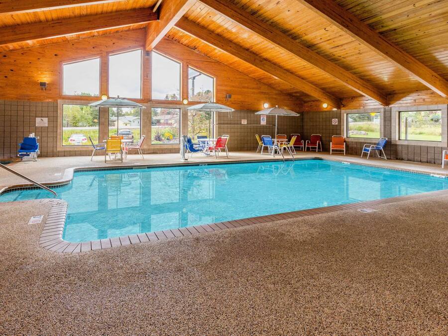 An indoor swimming pool with a wooden ceiling and tiled walls, surrounded by various colored lounge chairs.