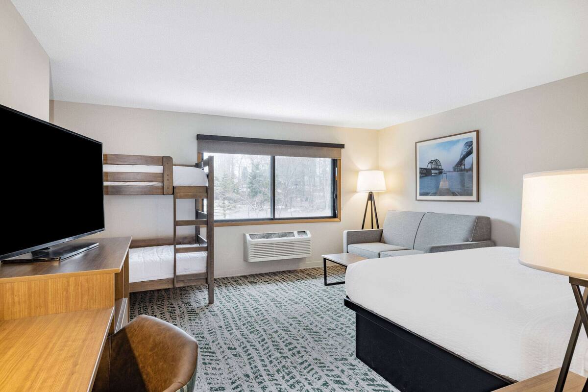 Een hotelkamer met een bed, een bureau met een televisie, een bank, een staande lamp en een raam met uitzicht op bomen.