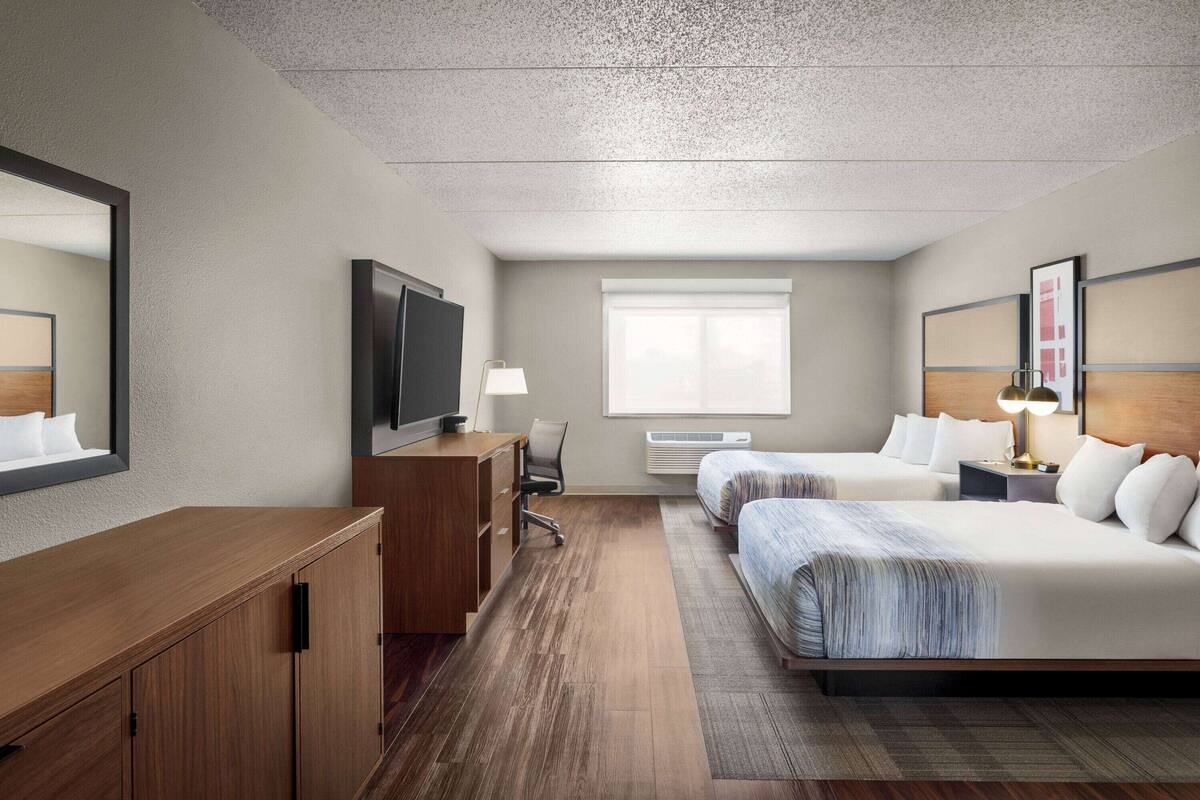 A hotel room with a wooden desk, a television, two beds, a window, and a mirror.