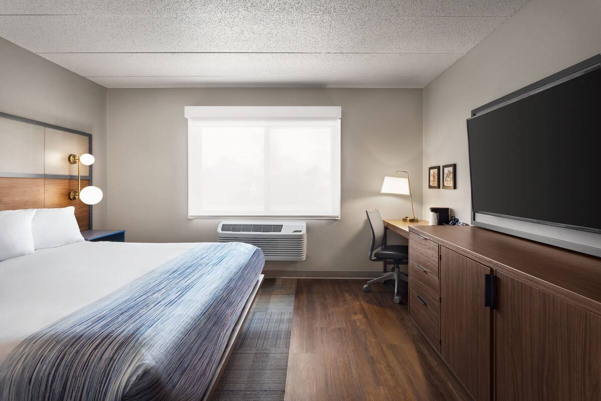 A hotel room with a bed, a desk, a chair, a large flat-screen TV, and a window with blinds.