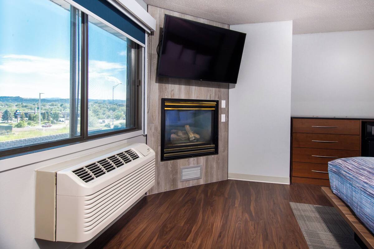 A room with a large window, a wall-mounted TV, a built-in fireplace, and a bed with a patterned bedspread.