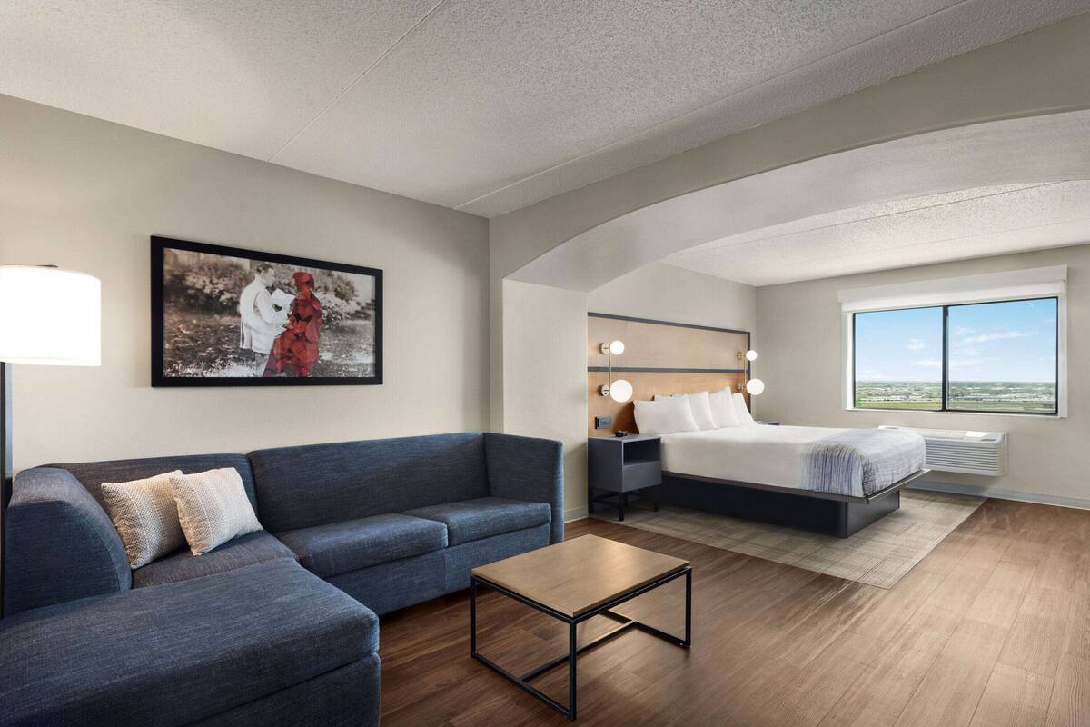 A modern hotel room with a sofa, a bed, a bedside table, a lamp, and a large window offering a view.