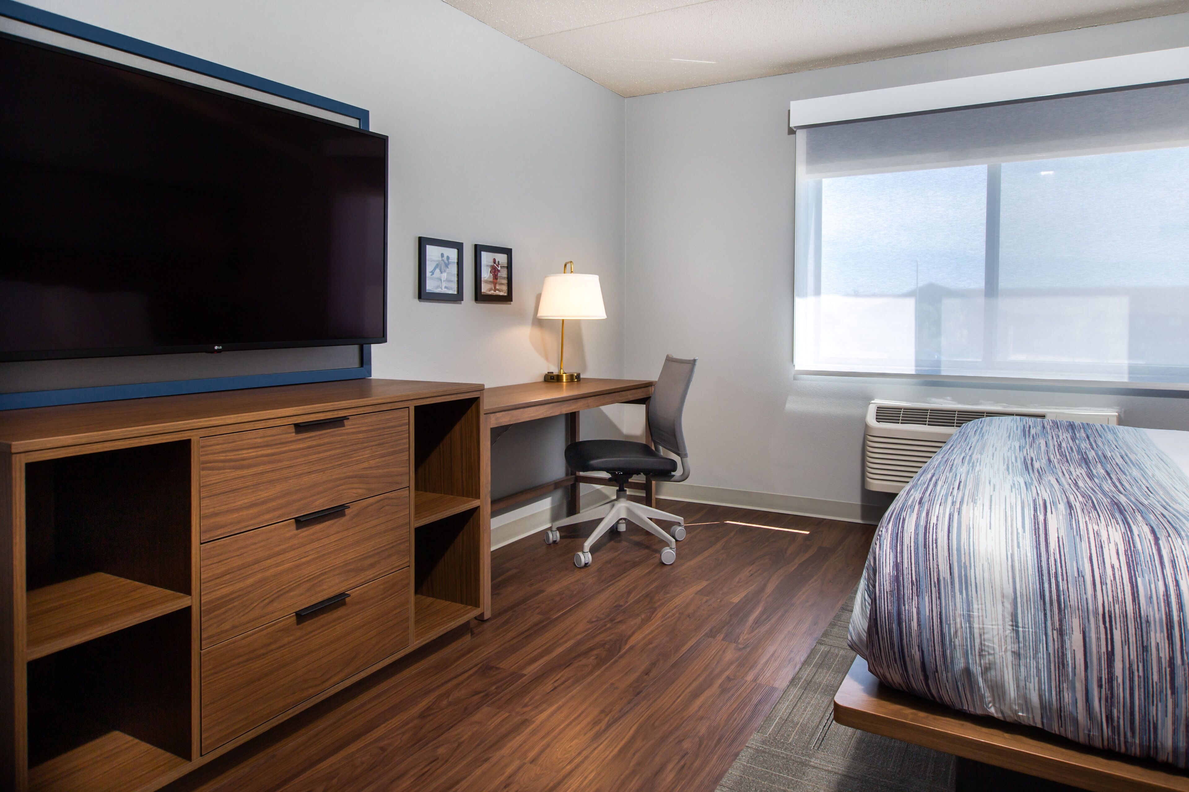 Een hotelkamer met een houten bureau, een stoel, een bed met een gestreept kleed, een televisie, een lamp en een raam met zonwering.