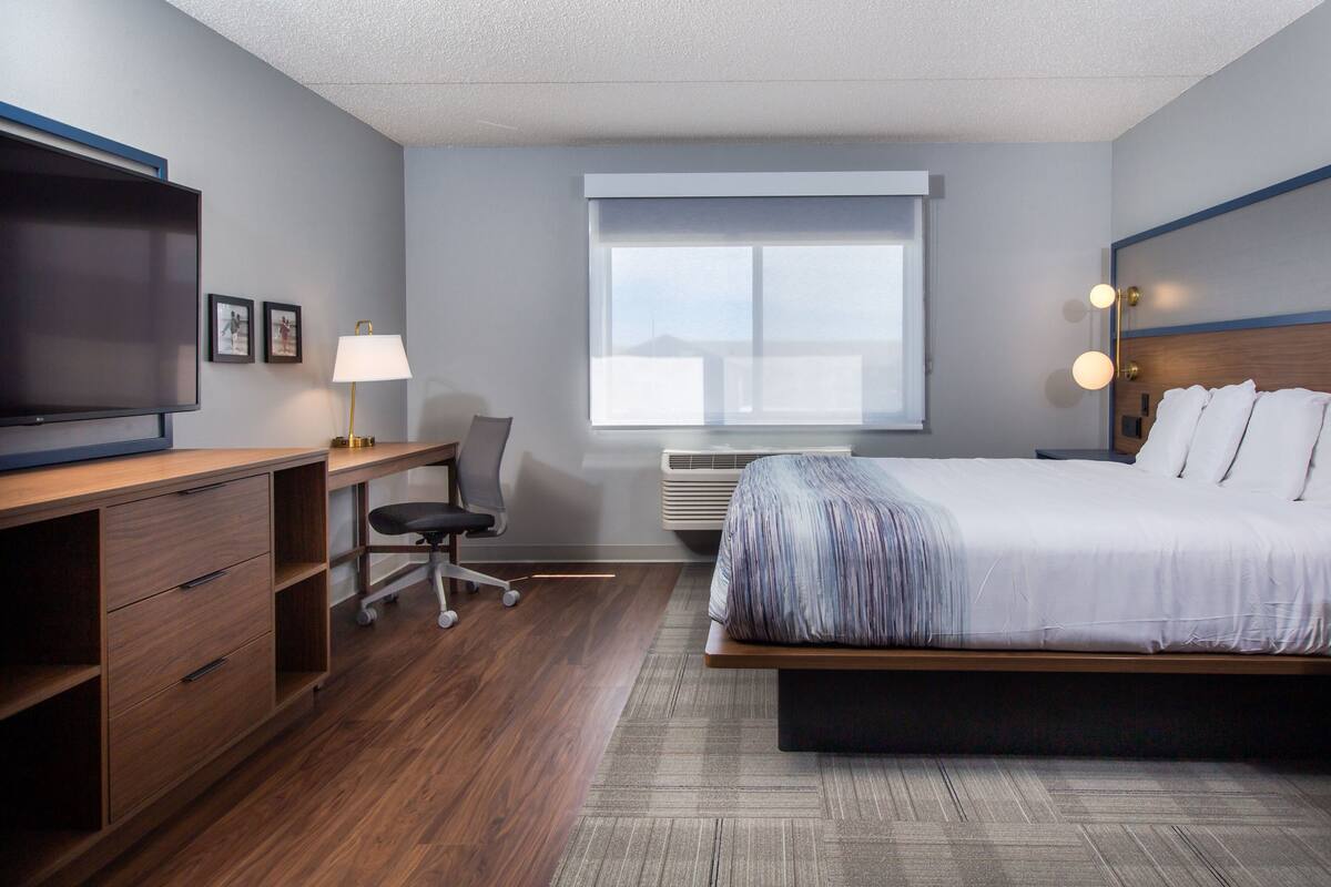 A modern hotel room with a large bed, a desk with a chair, a television, and a window with blinds.