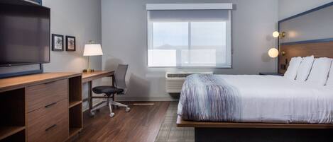 A modern hotel room with a large bed, a desk with a chair, a television, and a window with blinds.
