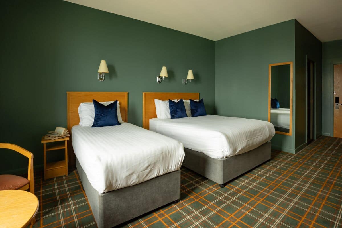 Quarto de hotel com duas camas, uma mesinha, uma cadeira e um espelho.