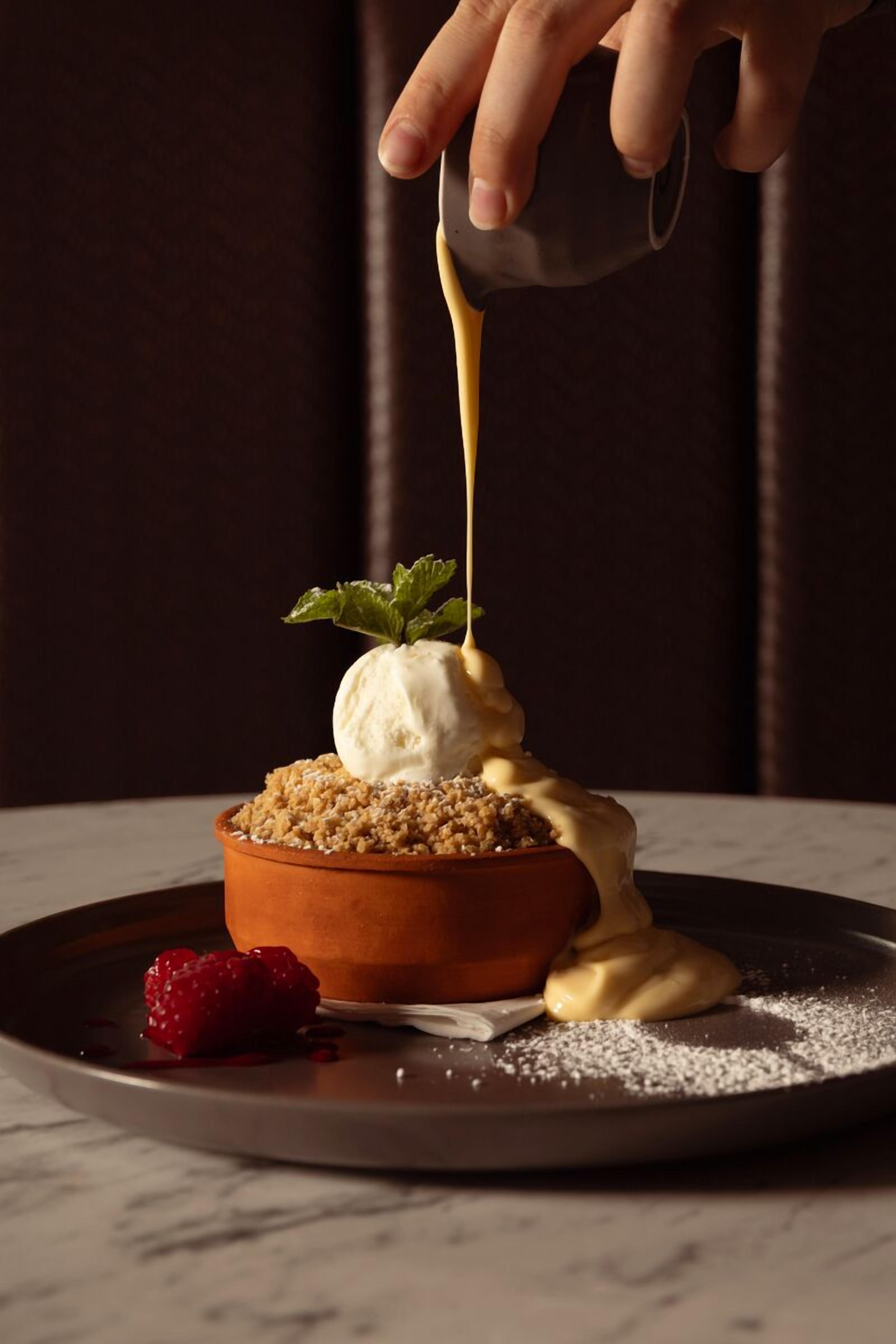 Ein Dessert mit einer Kugel Eis auf zerbröseltem Kuchen, das mit Soße beträufelt ist, und einer Himbeere daneben.