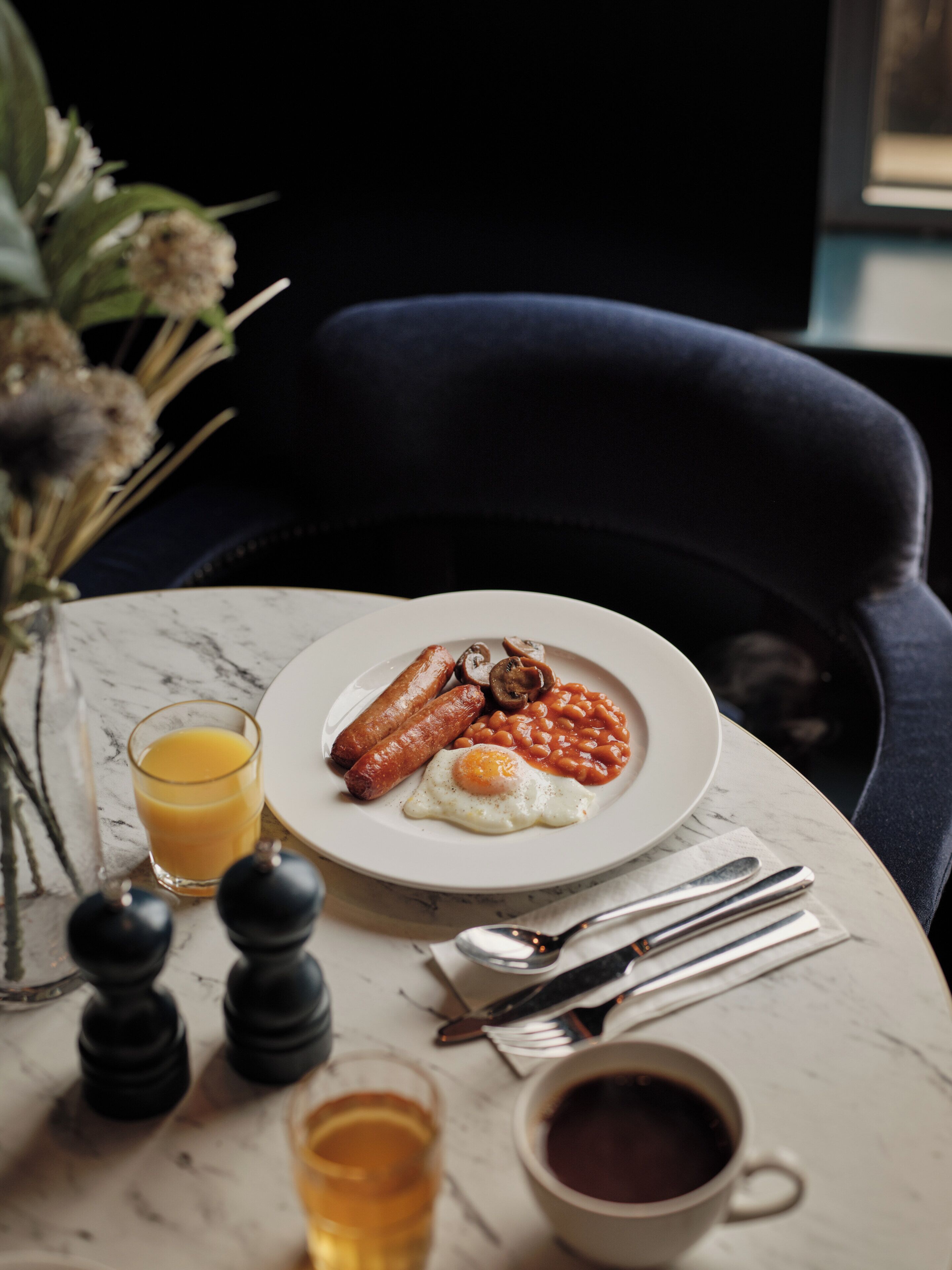 A breakfast plate with eggs, sausages, and beans, a glass of orange juice, and a cup of coffee on a marble table.