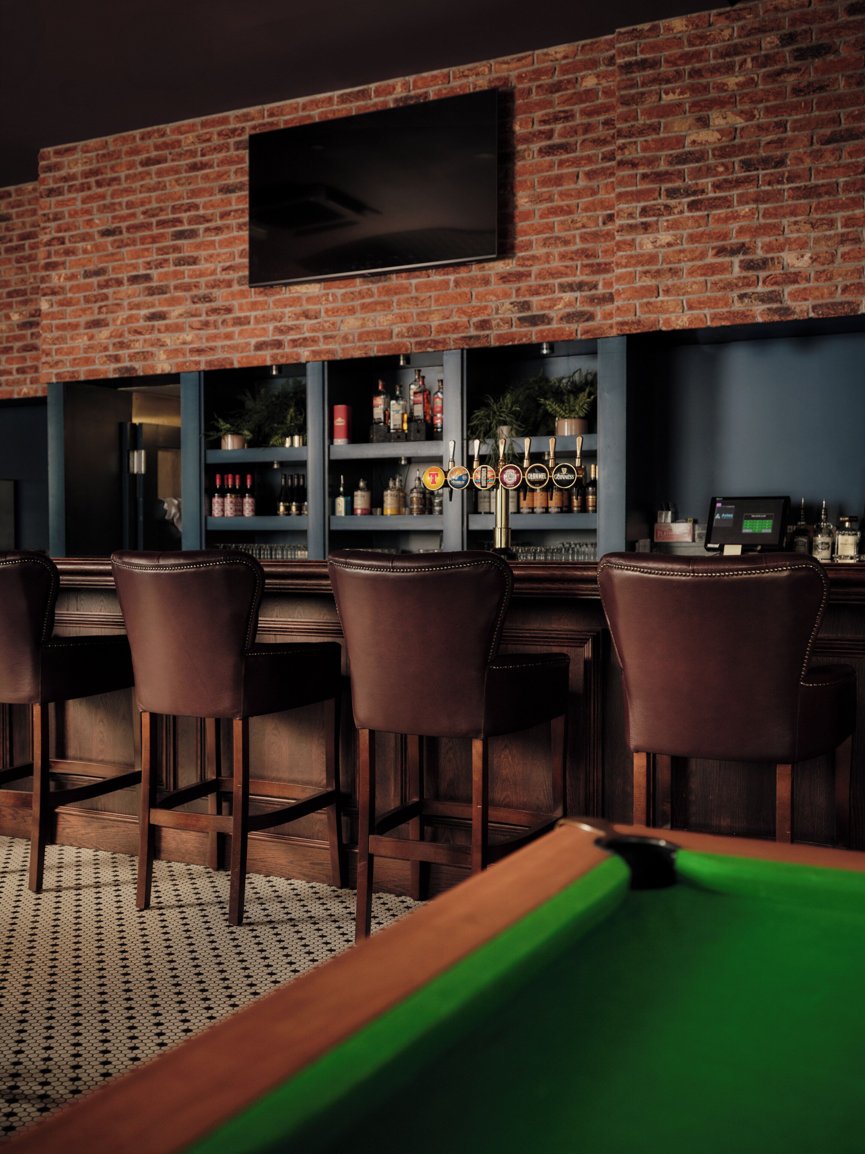 Un espace bar avec un mur en briques, un téléviseur fixé au mur et un comptoir de bar doté de plusieurs robinets et d’étagères remplies de bouteilles.