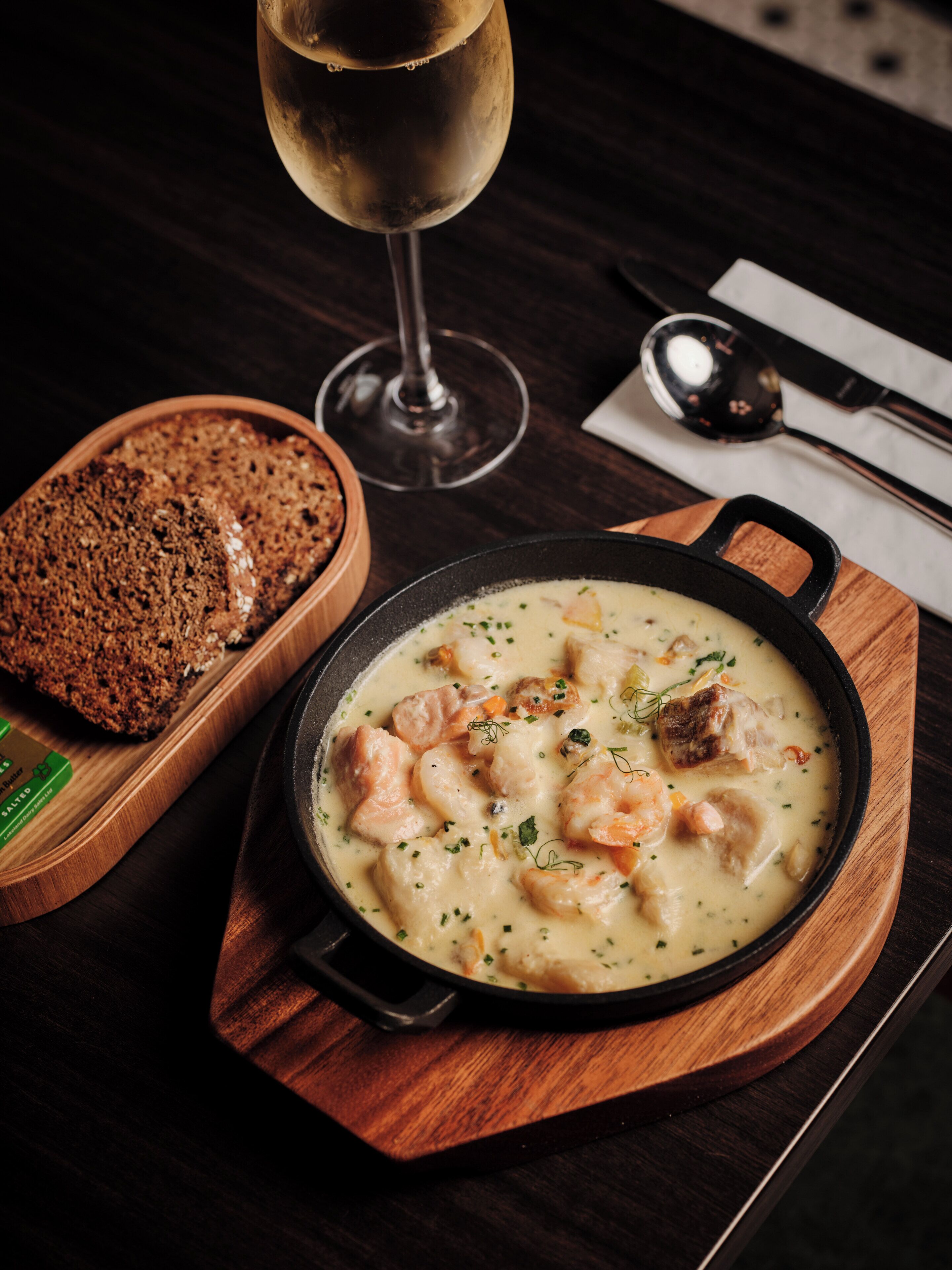 A wooden serving board with a bowl of creamy seafood stew, a glass of white wine, and a loaf of bread.