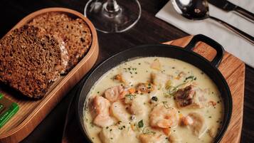 A wooden serving board with a bowl of creamy seafood stew, a glass of white wine, and a loaf of bread.