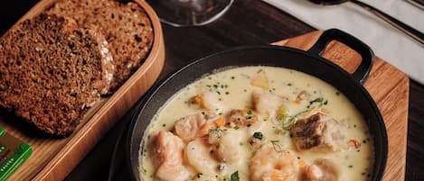 A wooden serving board with a bowl of creamy seafood stew, a glass of white wine, and a loaf of bread.