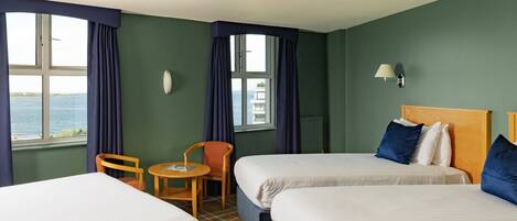 A hotel room with two beds, a small table, chairs, a lamp, and a view of the sea.