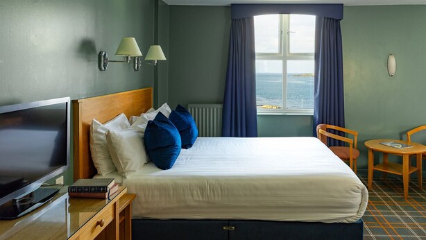A hotel room with a bed, a desk with a TV, a chair, and a view of the sea through a window.