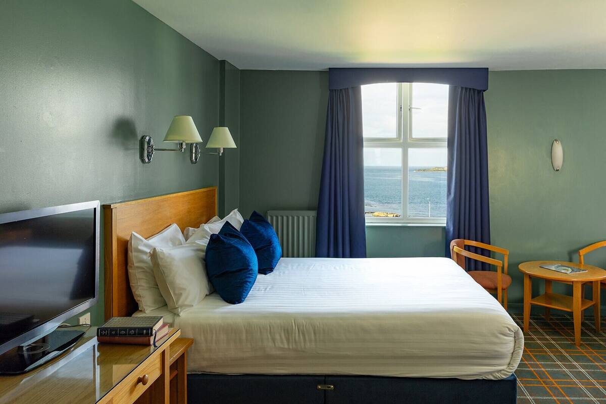 Quarto de hotel com cama, secretária com televisão, cadeira e vista para o mar através de uma janela.