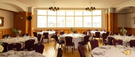 Un salón de banquetes con mesas redondas dispuestas para cenar, amplias ventanas que dejan entrar luz natural y candelabros colgando del techo.