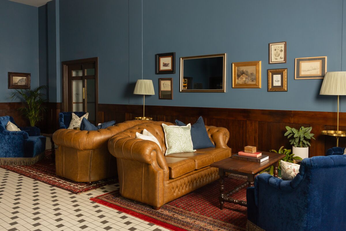 Une pièce avec un canapé en cuir marron, des fauteuils bleus, une table basse en bois et un tapis rouge à motifs.