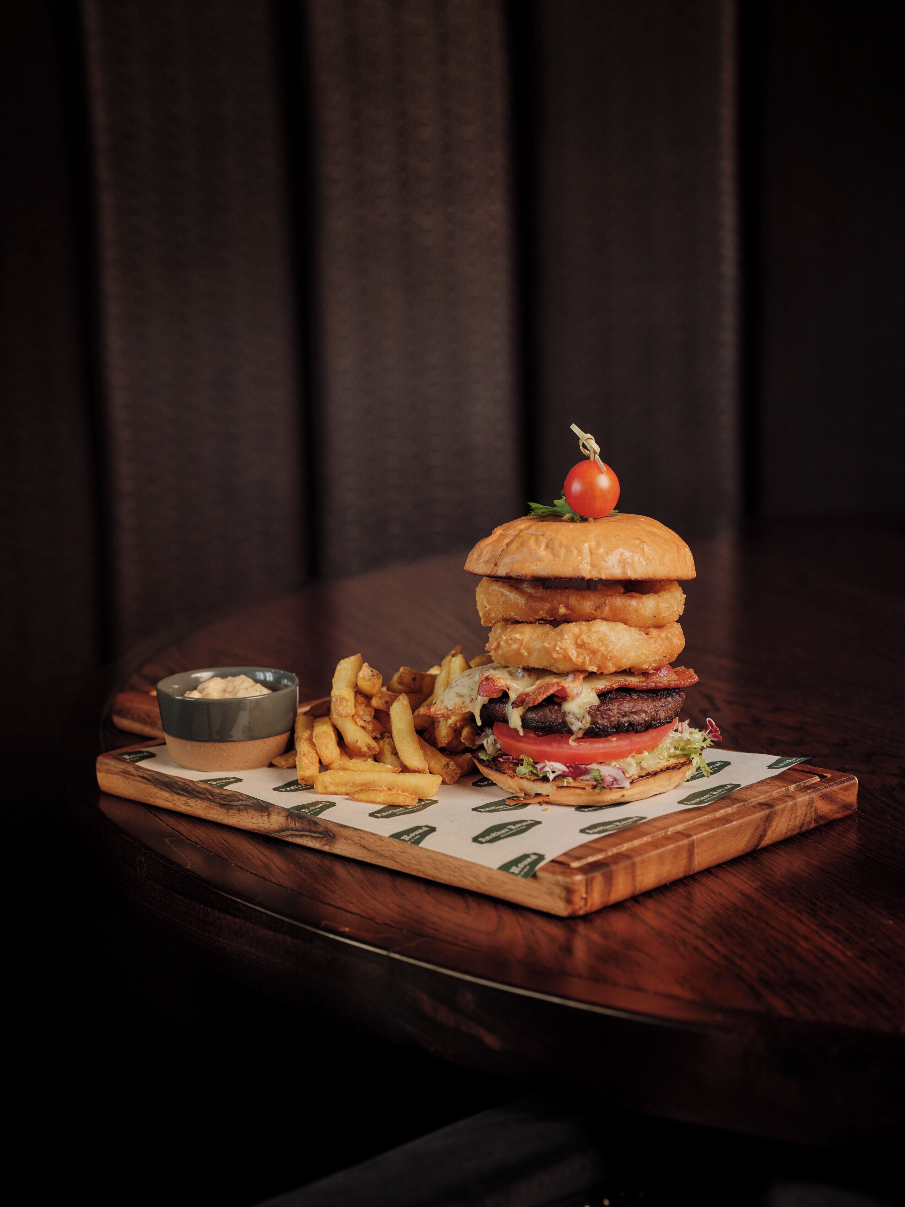 A burger with a tomato on top, fries, and a small bowl of sauce on a wooden board.