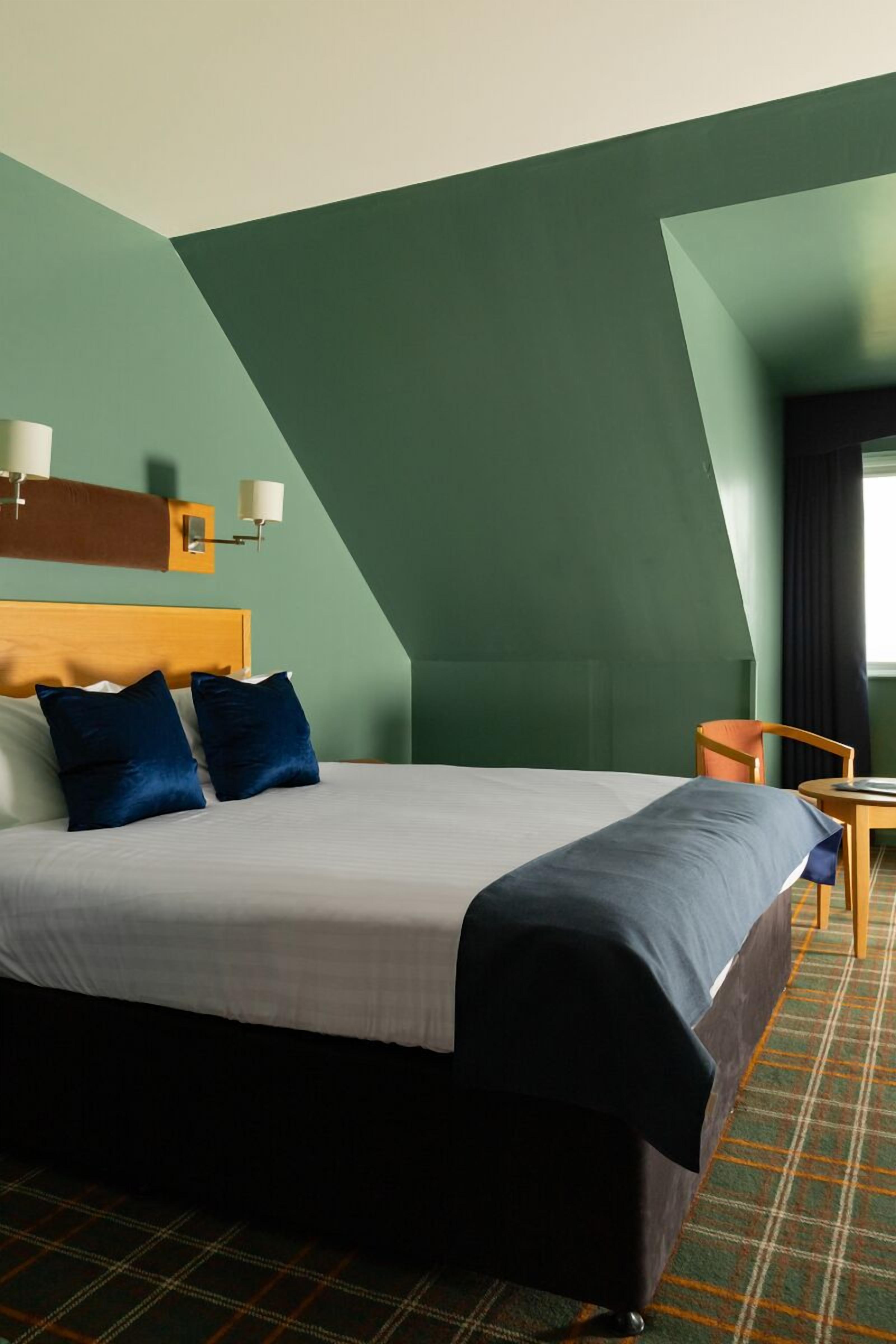 A hotel room with a large bed, a wooden headboard, two blue pillows, a plaid carpet, and a small table with a chair.