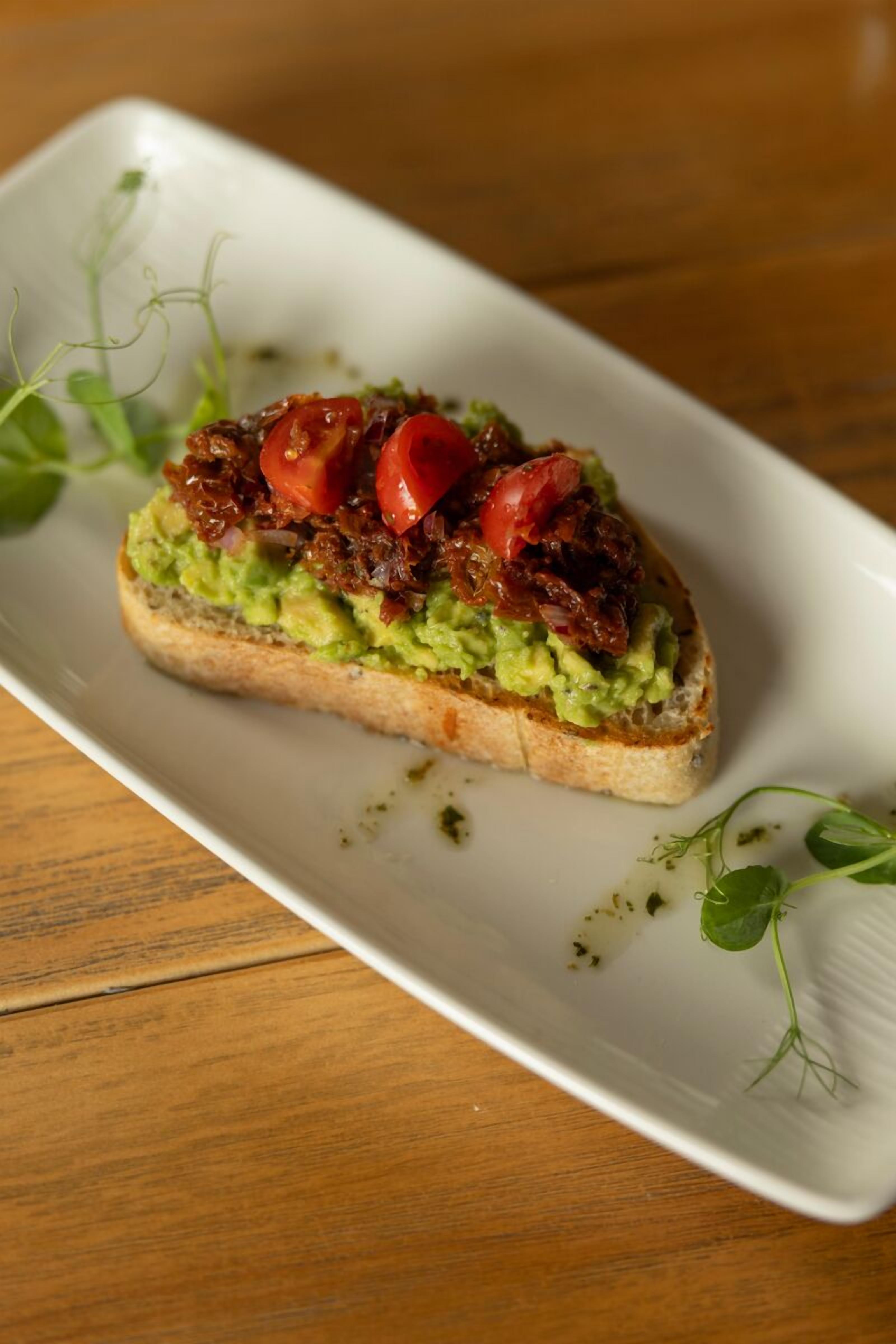 A rectangular white plate with a slice of toast topped with avocado and tomatoes, garnished with microgreens, on a wooden table.