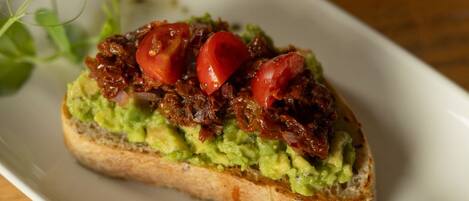 A rectangular white plate with a slice of toast topped with avocado and tomatoes, garnished with microgreens, on a wooden table.