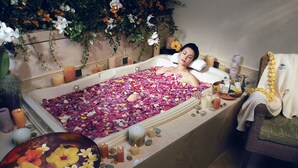 A bathtub filled with rose petals and candles, surrounded by toiletries and a wicker chair.
