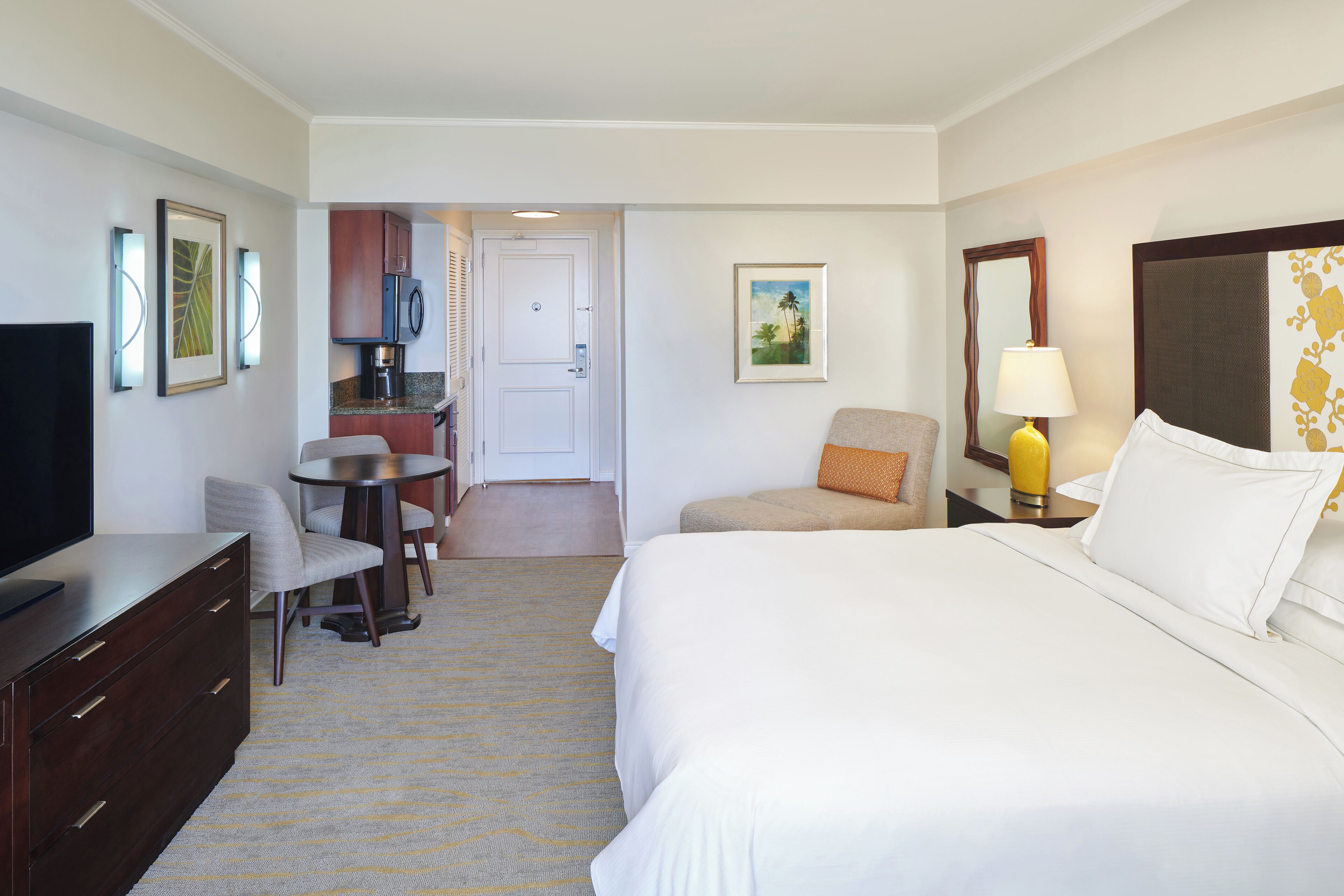 A hotel room with a bed, a desk, a chair, a TV, a lamp, and a painting on the wall.