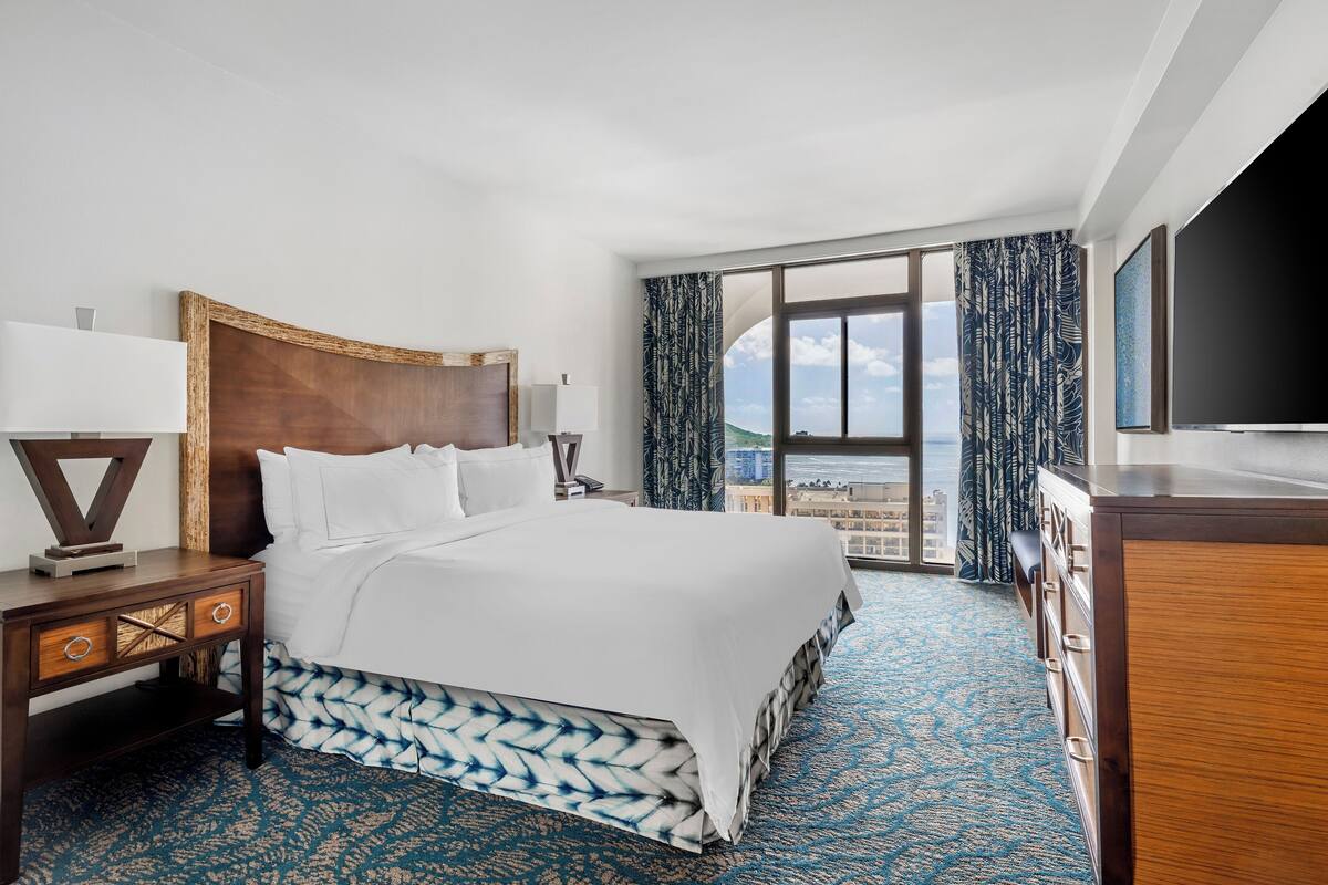 Ein Hotelzimmer mit einem großen Bett, einem Nachttisch aus Holz, einer Kommode und einem großen Fenster mit Blick auf die Stadt.