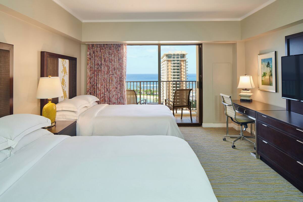 Ein Hotelzimmer mit zwei Betten, einem Schreibtisch, einem Stuhl, einem Fernseher und einem Balkon mit Blick auf das Meer und ein hohes Gebäude.