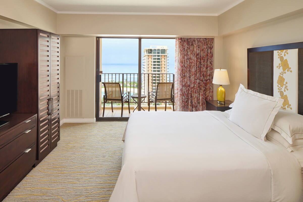Une chambre d’hôtel avec un grand lit, une télévision, une armoire, un balcon avec vue sur l’océan et un bâtiment, ainsi qu’une table avec des chaises.