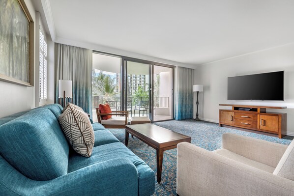 Une chambre d’hôtel avec un canapé bleu, un fauteuil beige, une table basse en bois, un téléviseur posé sur un meuble en bois, et une grande fenêtre donnant sur des bâtiments et des espaces verts.