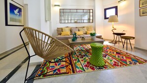 A modern living room with a sofa, a green ottoman, a wicker chair, a colorful rug, and a lamp. - Kalidon Beach Hotel (Samos)