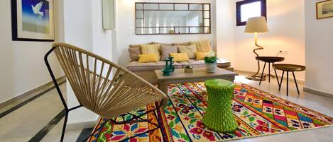 A modern living room with a sofa, a green ottoman, a wicker chair, a colorful rug, and a lamp.