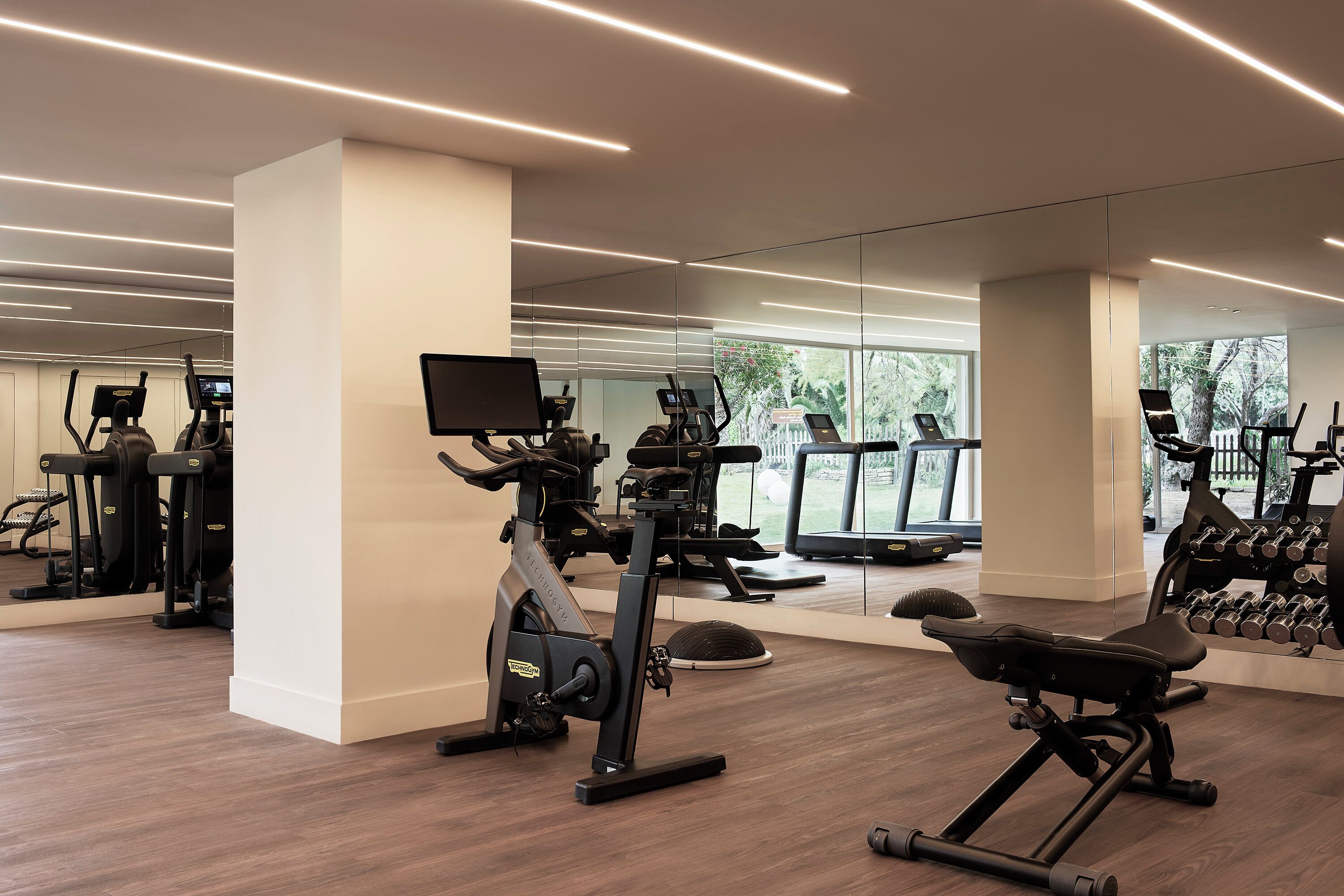 A modern gym with cardio machines, weight benches, and a large mirror reflecting the space.