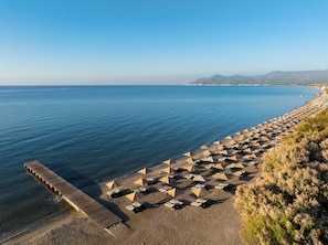 A beach with rows of sun loungers and umbrellas, a wooden pier, and a clear view of the sea and distant hills. - Doryssa Seaside Resort (Samos)