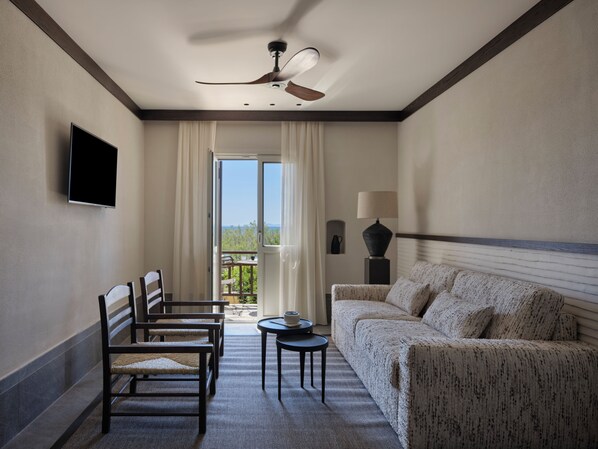 A hotel room with a sofa, chairs, a TV, a ceiling fan, a lamp, and a view through a door.