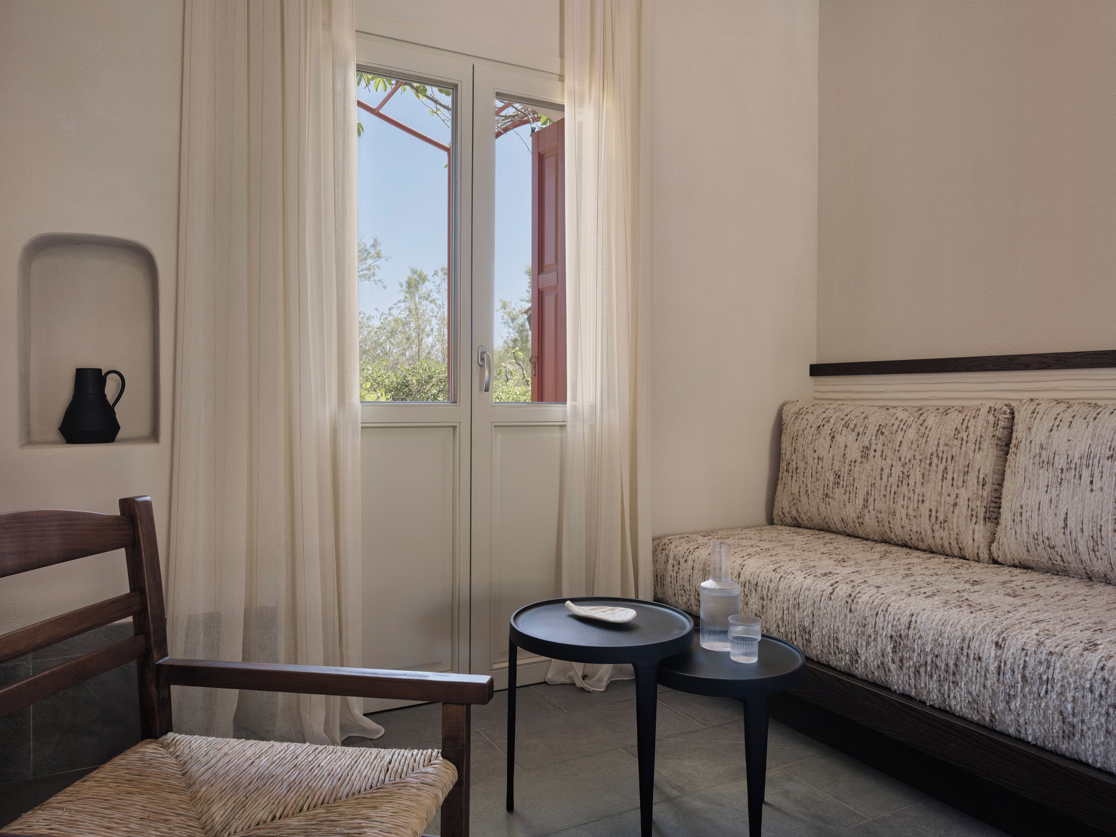 Une pièce comprenant une chaise en bois, une petite table ronde avec un verre, un canapé et une fenêtre avec des rideaux.