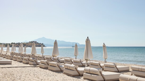 A beach with rows of beige lounge chairs and umbrellas, a clear sky, and a calm sea with a distant mountain. - Doryssa Seaside Resort (Samos)