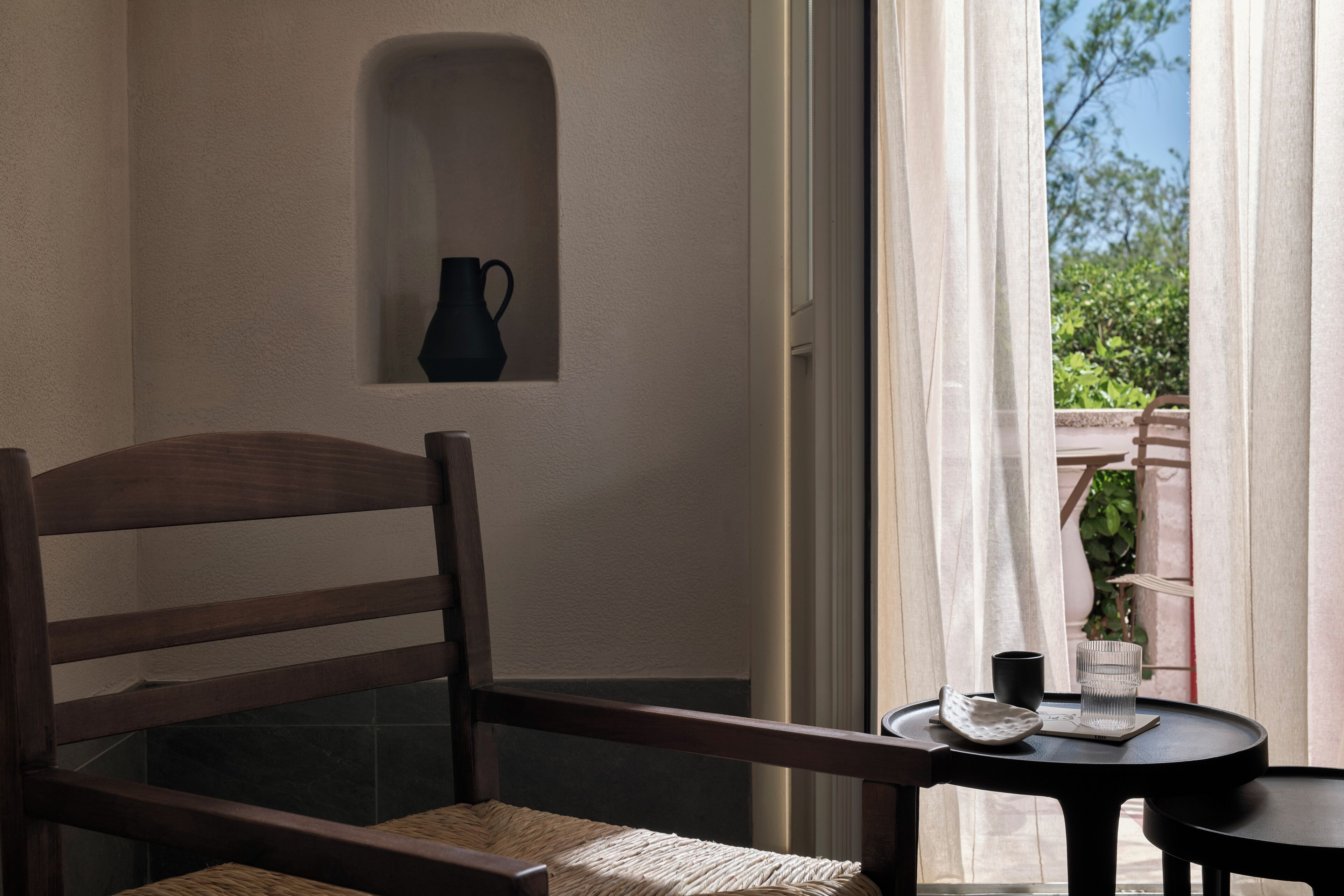Una silla y mesa de madera con una jarrón y taza, un pequeño estante con un jarrón, y vista a través de una ventana con cortinas.