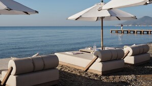 A beachside lounge with white umbrellas and beige lounge chairs, a wooden deck, and a view of the sea and a pier in the background. - Doryssa Seaside Resort (Samos)
