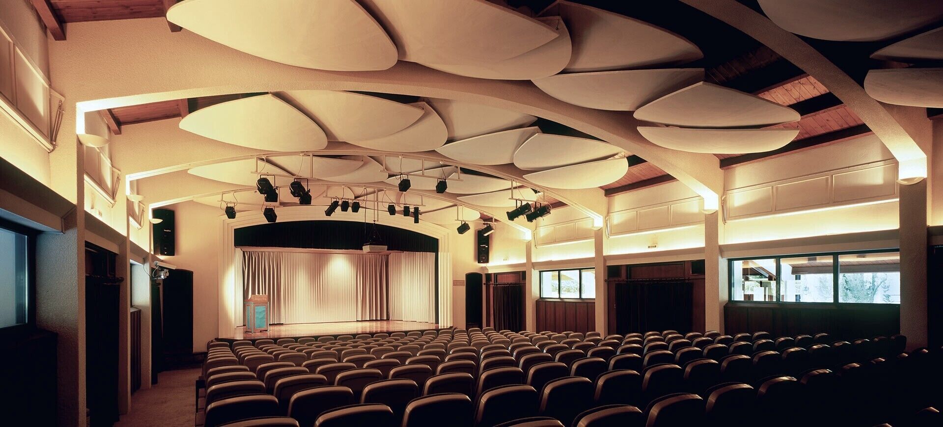 Ein Auditorium mit terrassenförmigen Sitzreihen, einer Bühne mit Vorhängen und einer Decke mit besonderen Leuchten.