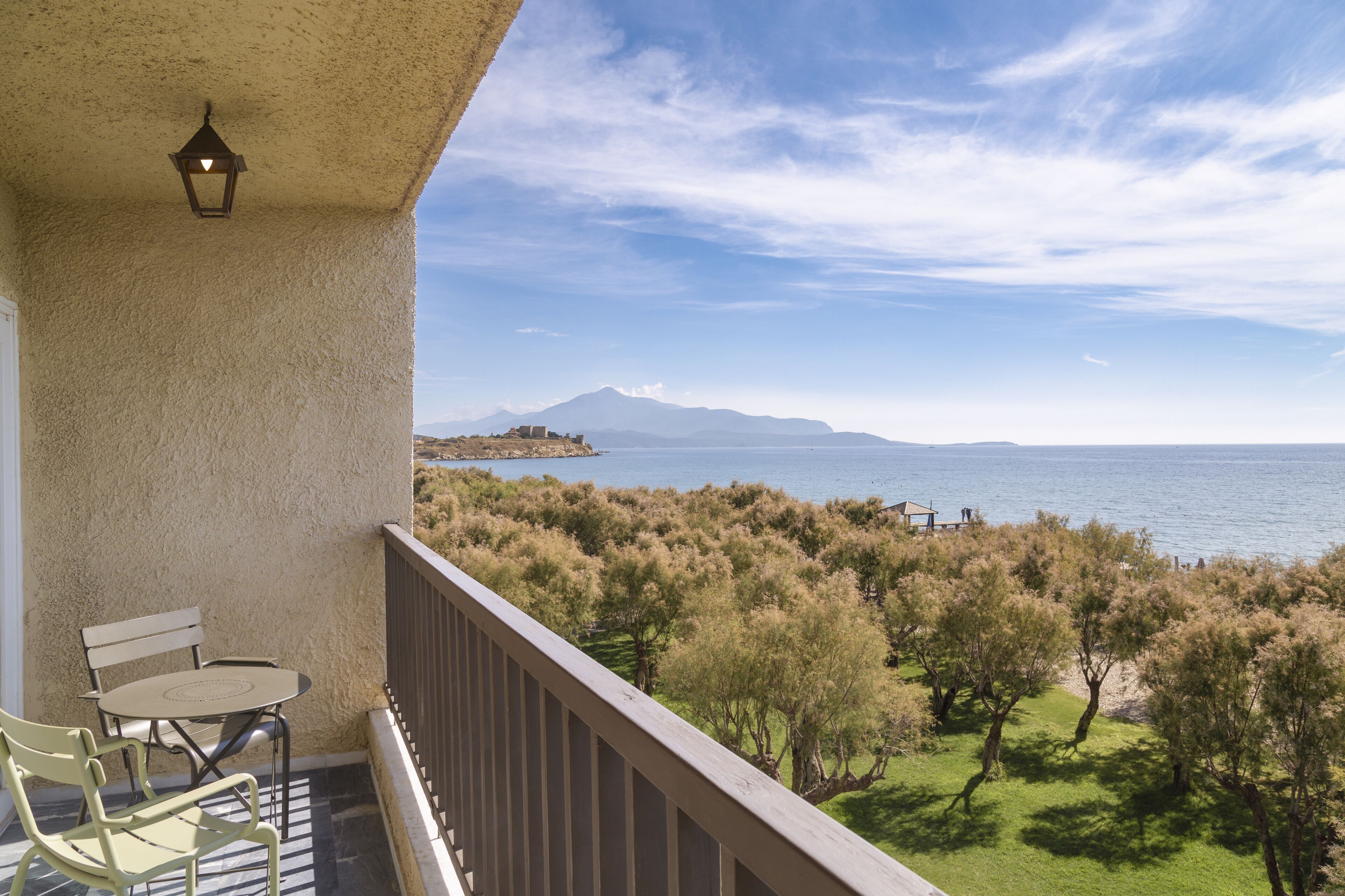A balcony with a table and chairs, overlooking a coastal landscape with trees and a distant mountain.