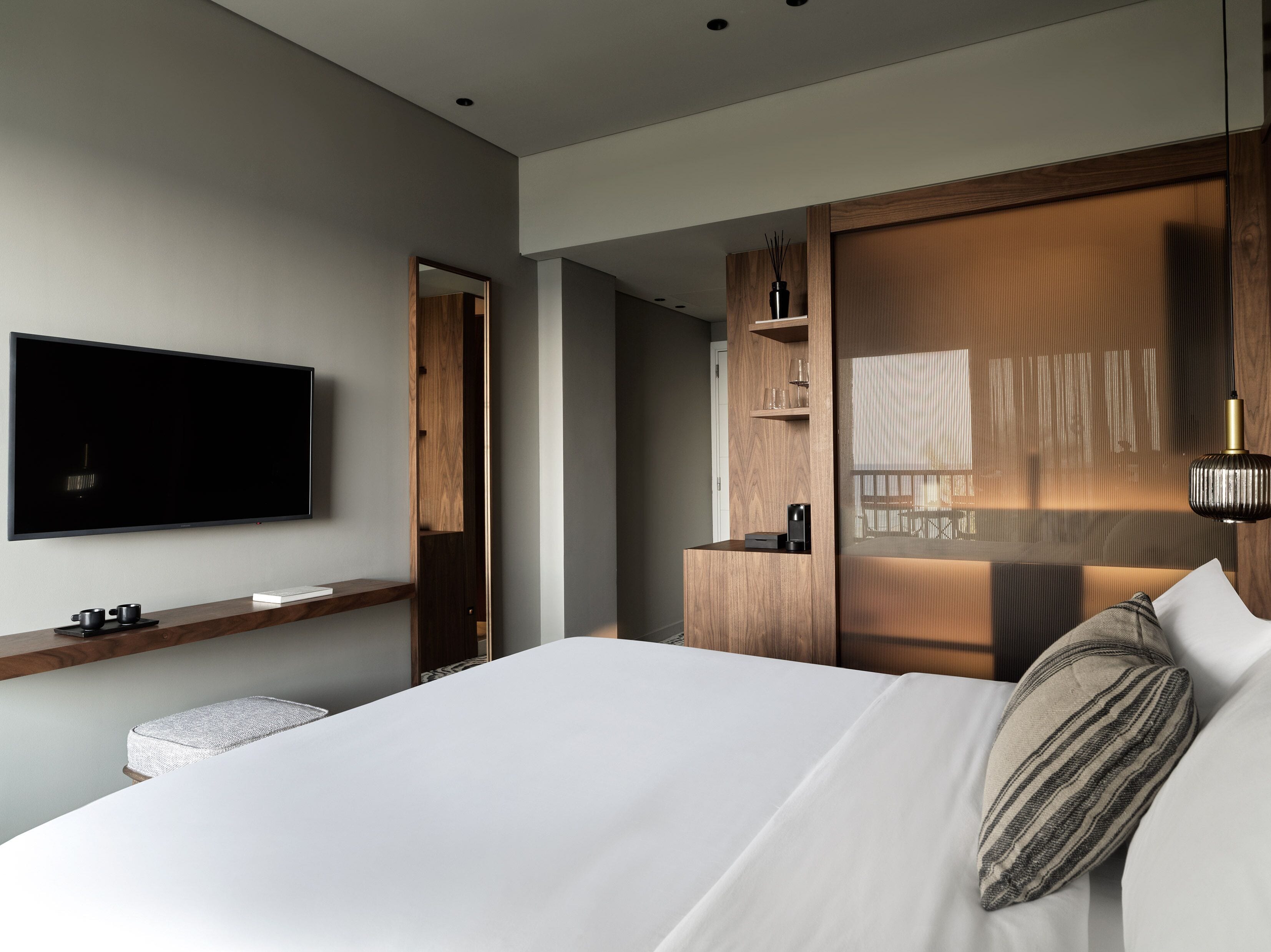 Una habitación de hotel moderna con una cama grande, un televisor de pantalla plana montado en la pared, una estantería de madera con una cafetera y un armario empotrado con espejo.