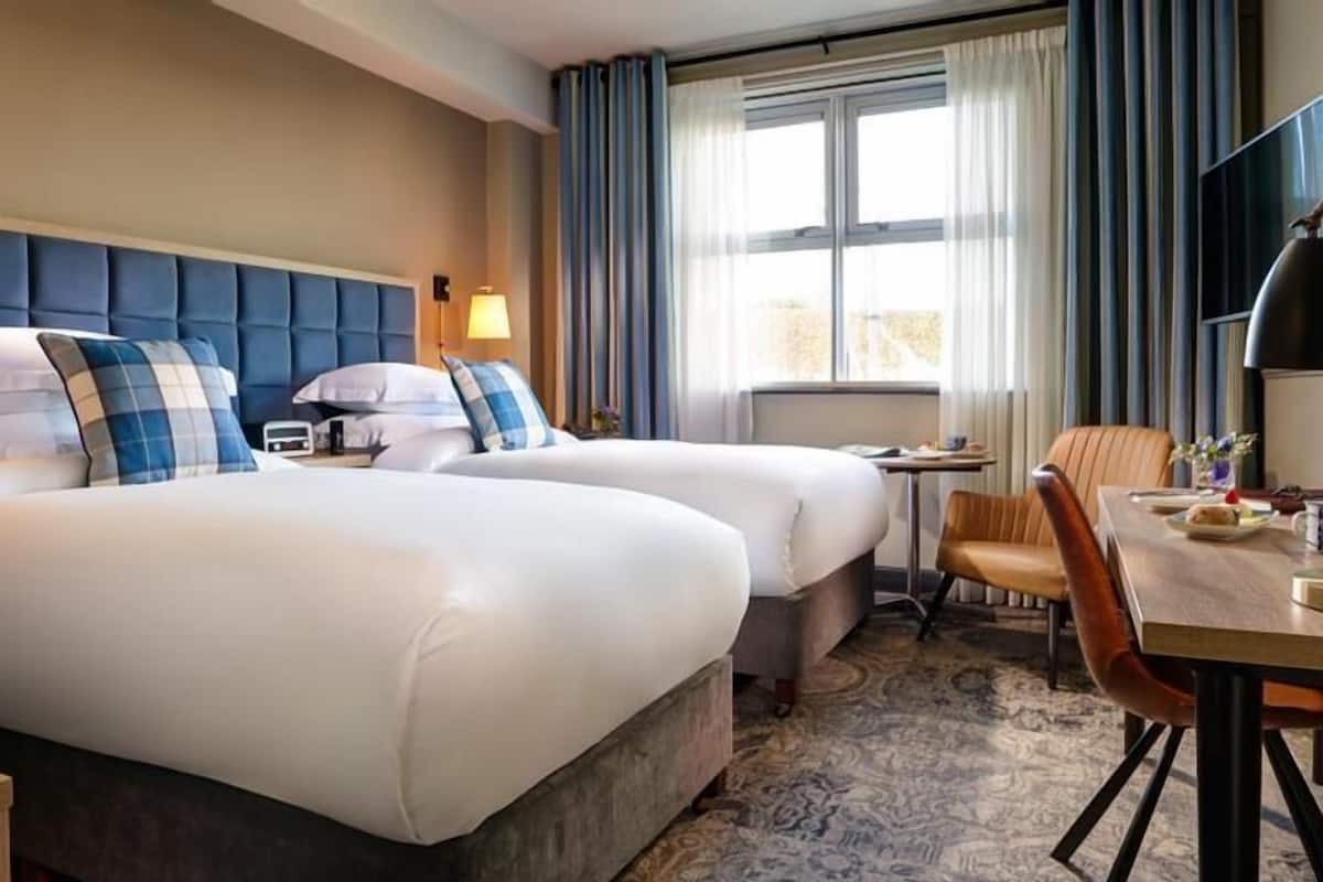 A hotel room with two beds, a desk, a chair, a TV, and a window with curtains.