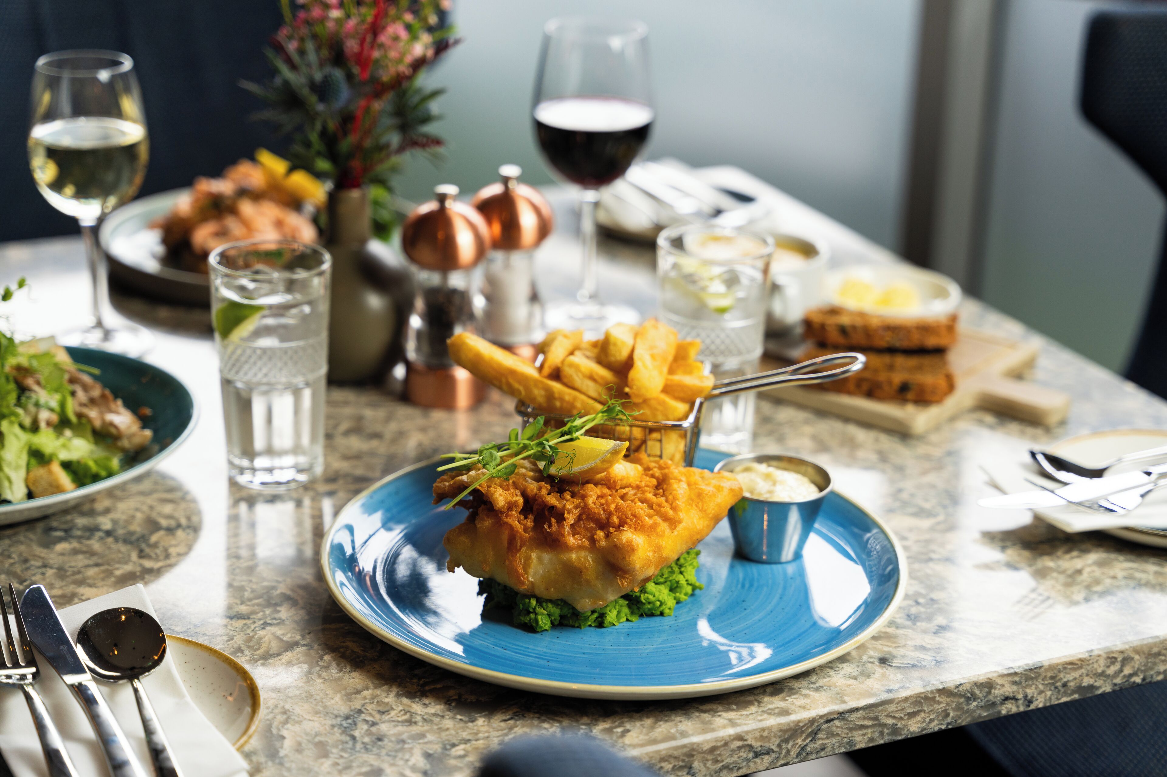 Una mesa de comedor con un plato de pescado y papas fritas, una copa de vino blanco y una copa de vino tinto.