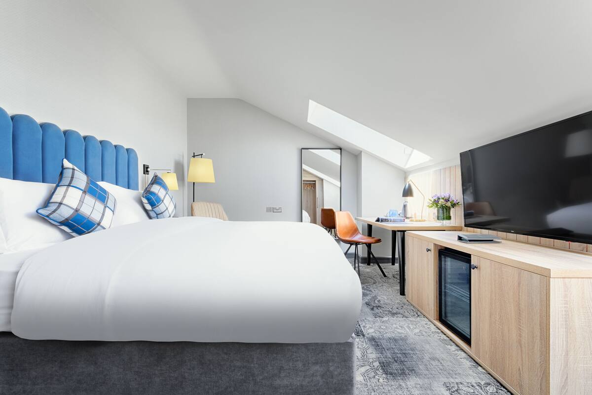 A modern hotel room with a large bed, a desk with a flat-screen TV, a mini-fridge, and a seating area with chairs.