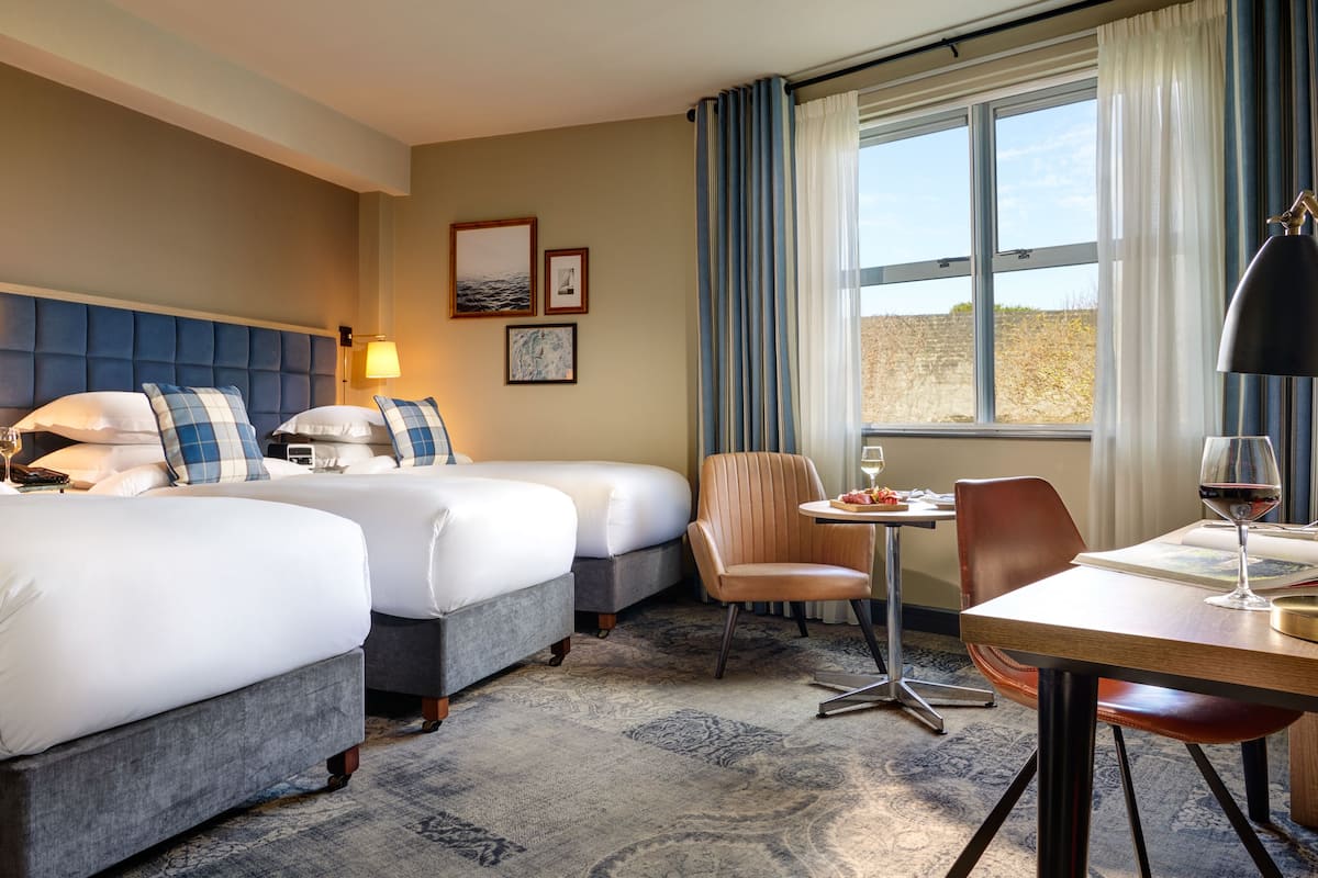 A hotel room with two beds, a dining table, a chair, a lamp, and a window with curtains.