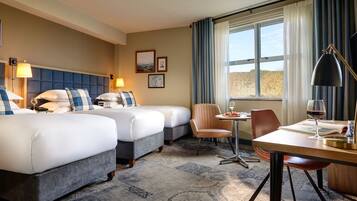 A hotel room with two beds, a dining table, a chair, a lamp, and a window with curtains.